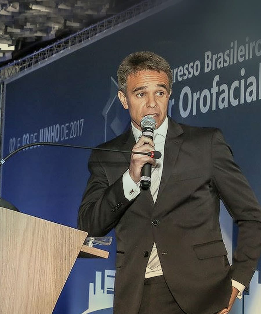 Dr. Rodrigo Wendel, cirurgião-dentista, falando ao microfone em um congresso, diante do painel azul do evento.